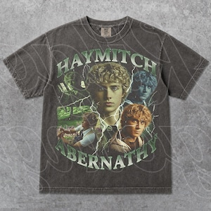 May include: A charcoal grey short-sleeve t-shirt featuring a graphic print of Haymitch Abernathy. The design includes multiple portraits of the character, green lightning bolt accents, and the text HAYMITCH ABERNATHY in a bold, arched green font.