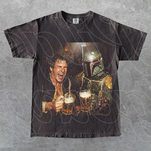 May include: A charcoal gray short-sleeve t-shirt featuring a graphic print of a smiling man and a character in a green and silver helmet holding glass beer mugs. The shirt has a crew neckline and a faded, vintage-style wash throughout the fabric.