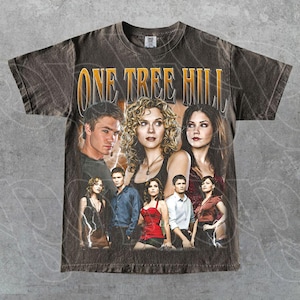 May include: A dark gray t-shirt featuring a vintage-style design with the text "ONE TREE HILL" in gold lettering. The shirt displays a collage of actors and actresses from the television show, with a distressed, faded appearance.