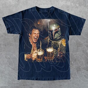 May include: A dark blue t-shirt featuring a graphic print of a laughing man and a character in a green and brown helmeted suit, both holding glass beer mugs. The design shows the two figures in a dimly lit setting, clinking their drinks together.