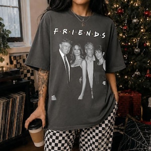 May include: A charcoal gray short-sleeve t-shirt featuring a black and white graphic print of four individuals standing together. Above the figures, the word "F R I E N D S" is printed in a white serif font. The shirt has a crew neckline and a relaxed fit.