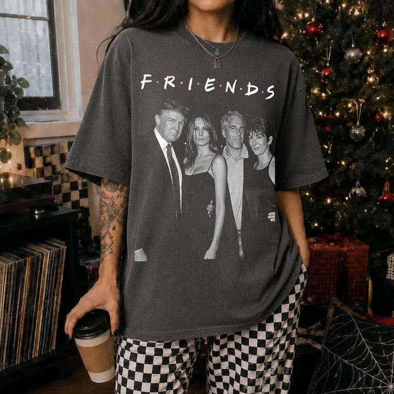 May include: A charcoal gray short-sleeve t-shirt featuring a black and white graphic print of four individuals standing together. Above the figures, the word "F R I E N D S" is printed in a white serif font. The shirt has a crew neckline and a relaxed fit.