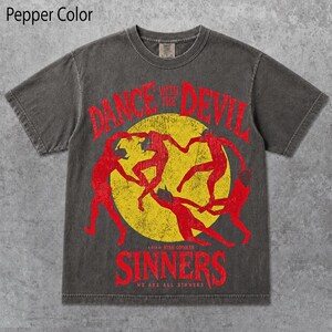 Può includere: Una t-shirt a maniche corte color pepe con una grande grafica a cerchio giallo e sagome rosse di figure danzanti. Il testo DANCE WITH THE DEVIL è scritto in rosso sopra il cerchio e SINNERS in rosso sotto, con del testo più piccolo incluso.