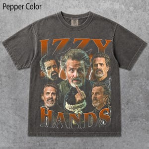 May include: A pepper-colored short-sleeve t-shirt featuring a graphic print of a man with a beard in four different poses. The central figure is shown with hands bound by rope, gesturing with a finger. The text IZZY HANDS is printed in orange.