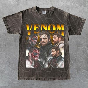 Puede incluir: Camiseta gris oscuro con la palabra "VENOM" en dorado y un collage de imágenes. Las imágenes muestran a un hombre con un parche en el ojo y varias expresiones faciales. La camiseta tiene un aspecto vintage y desgastado.