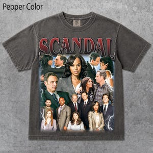 May include: A pepper-coloured short-sleeve t-shirt featuring a graphic print of the cast of the television show Scandal. The word SCANDAL is printed in large red and white letters at the top, with various characters arranged below against a blue background.