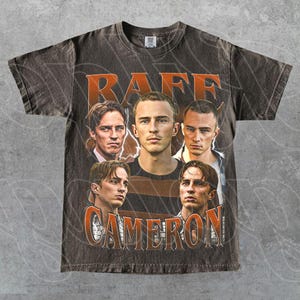 May include: A charcoal gray short-sleeve t-shirt featuring a graphic print of four portraits of the same individual. The text "RAFF" appears in large orange letters at the top, and the text "CAMERON" appears in large orange letters at the bottom.