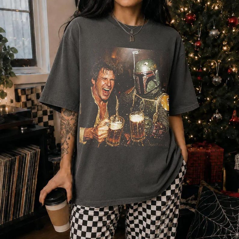 May include: A charcoal gray short-sleeve t-shirt featuring a graphic print of a man and a helmeted character clinking beer mugs together. The shirt has a crew neckline and a relaxed fit, showcasing a detailed, colorful illustration on the front.