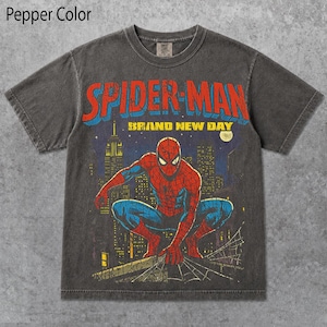 May include: A charcoal gray short-sleeve t-shirt featuring a graphic of Spider-Man crouching on a rooftop. The text reads SPIDER-MAN and BRAND NEW DAY. The design includes a city skyline background with a yellow moon, rendered in red, blue, and yellow.