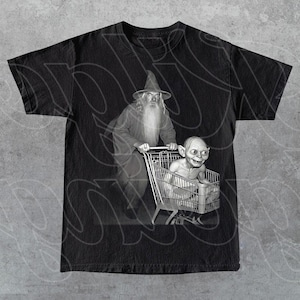 May include: Black t-shirt featuring a grayscale image of a wizard pushing a shopping cart with Gollum inside. The wizard wears a tall hat and long robe. Gollum is seated in the cart, smiling. The design is printed on a black t-shirt.