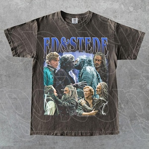 Puede incluir: Camiseta marrón oscuro con un diseño gráfico que presenta el texto "ED & STEDE" en azul. El diseño muestra varias figuras en una escena con relámpagos. La camiseta está hecha de un material suave.