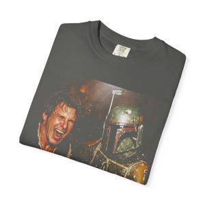 May include: A charcoal gray t-shirt featuring a graphic print of a laughing man with light brown hair next to a green and brown armored character wearing a helmet with a T-shaped visor. The shirt has a Comfort Colors brand label at the neck.
