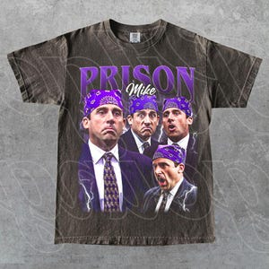 May include: Dark gray t-shirt featuring a graphic of the character "Prison Mike" from The Office. The design includes multiple images of the character wearing a purple bandana, with the words "Prison Mike" in purple text.