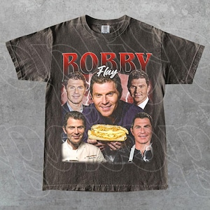 May include: Dark gray t-shirt featuring a collage of images of Bobby Flay, with the name "Bobby Flay" in red lettering. The shirt includes images of Bobby Flay holding a sandwich. The shirt has a vintage, distressed look.
