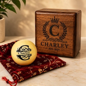 Peut inclure: Une boîte en bois brun foncé gravée d'une couronne, d'une couronne de laurier, de la lettre C et du texte CHARLEY EST. 2024. À côté se trouve une sphère crème avec un C noir et le nom Charley, posée sur une pochette en velours rouge.
