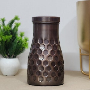 Puede incluir: Un jarr&oacute;n decorativo de color bronce con tapa. El jarr&oacute;n tiene un patr&oacute;n de panal en el cuerpo y un acabado met&aacute;lico liso. El jarr&oacute;n es cil&iacute;ndrico con una base ligeramente m&aacute;s ancha y una parte superior m&aacute;s estrecha.