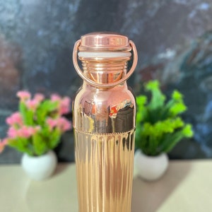 May include: A copper water bottle with a ribbed design and a copper lid with a white seal. The bottle has a handle on the lid.