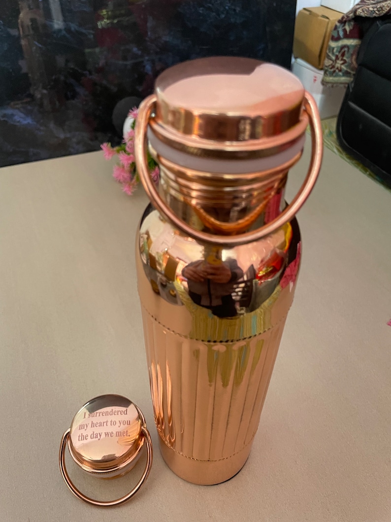 May include: A rose gold water bottle with a matching lid and a handle. The lid has an engraved message that reads "I surrendered my heart to you the day we met."