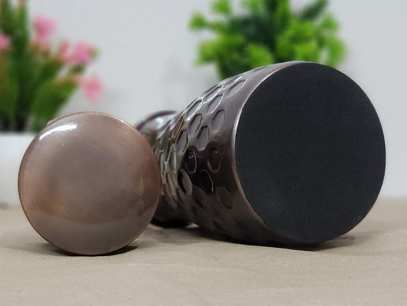 Puede incluir: Un tap&oacute;n de botella de color bronce con una textura martillada en el cuerpo y una base redonda y plana. El tap&oacute;n est&aacute; junto a una tapa redonda a juego. El fondo est&aacute; borroso, con toques de verde y rosa.