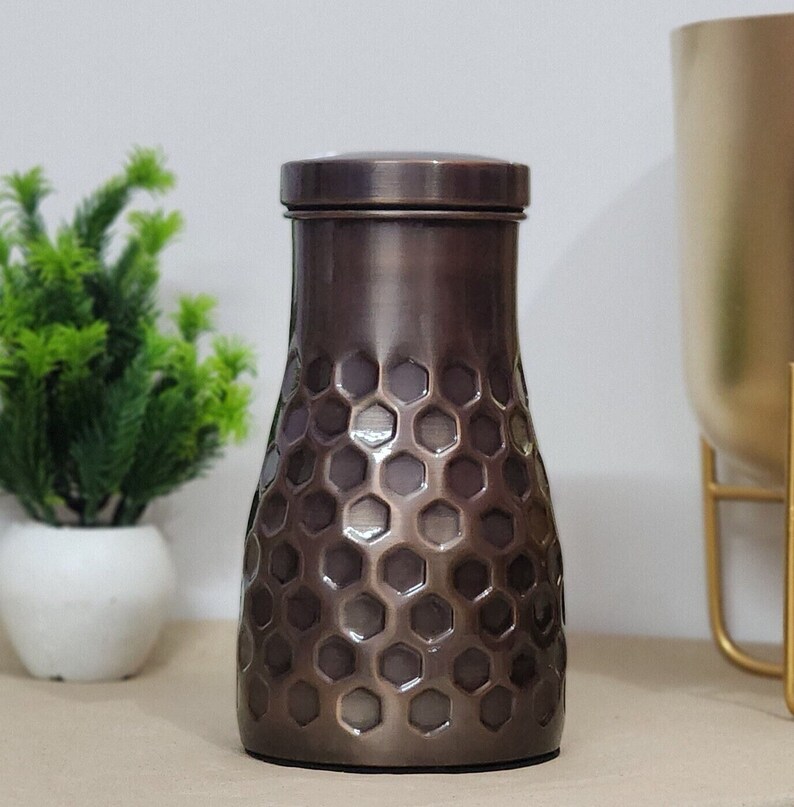 Puede incluir: Un jarr&oacute;n decorativo de color bronce con tapa. El jarr&oacute;n tiene un patr&oacute;n de panal en el cuerpo y un acabado met&aacute;lico liso. El jarr&oacute;n es cil&iacute;ndrico con una base ligeramente m&aacute;s ancha y una parte superior m&aacute;s estrecha.