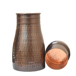 Puede incluir: Una jarra de agua de color cobre con una textura martillada y una tapa a juego. La jarra tiene una base ancha que se estrecha hacia arriba, con una banda decorativa cerca de la parte superior. La tapa es redonda y est&aacute; al lado de la jarra. El fondo es blanco.