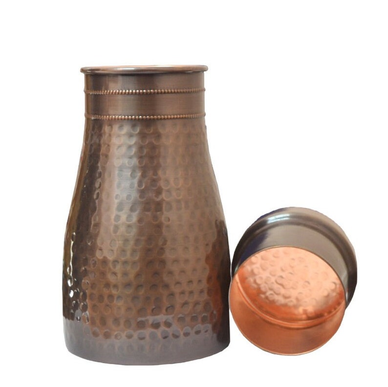 Puede incluir: Una jarra de agua de color cobre con una textura martillada y una tapa a juego. La jarra tiene una base ancha que se estrecha hacia arriba, con una banda decorativa cerca de la parte superior. La tapa es redonda y est&aacute; al lado de la jarra. El fondo es blanco.