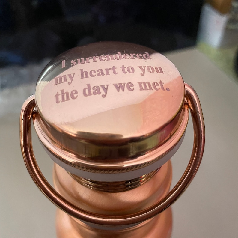 May include: A copper water bottle with a copper lid and a handle. The lid has an inscription that reads "I surrendered my heart to you the day we met."