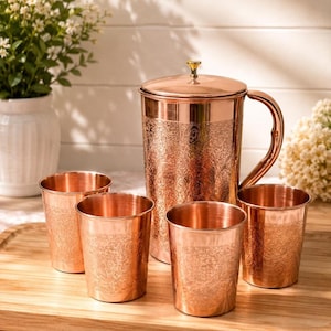 May include: A copper pitcher with a lid and handle, accompanied by four matching copper tumblers. Each piece features an intricate, embossed floral pattern covering the exterior surface, showcasing a polished metallic finish throughout.