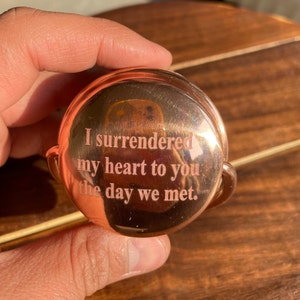 May include: A copper-colored round trinket with a hinged lid. The lid is engraved with the words "I surrendered my heart to you the day we met."