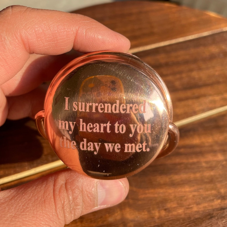 May include: A copper-colored round trinket with a hinged lid. The lid is engraved with the words "I surrendered my heart to you the day we met."