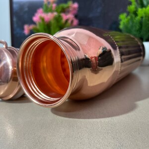 May include: A copper water bottle with a lid. The bottle has a shiny, polished finish and is designed with vertical ridges. The bottle is open and the inside is visible.