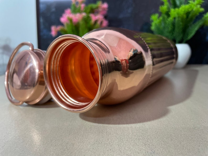 May include: A copper water bottle with a lid. The bottle has a shiny, polished finish and is designed with vertical ridges. The bottle is open and the inside is visible.