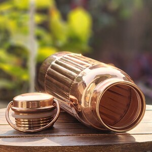 May include: A copper water bottle with a ribbed design and a matching lid. The bottle is open and the inside is visible.