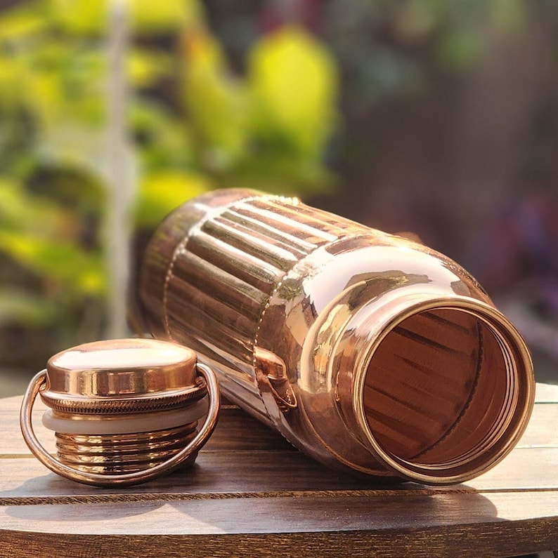 May include: A copper water bottle with a ribbed design and a matching lid. The bottle is open and the inside is visible.