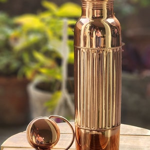 May include: A copper water bottle with a ribbed design and a matching lid. The bottle is on a wooden surface.