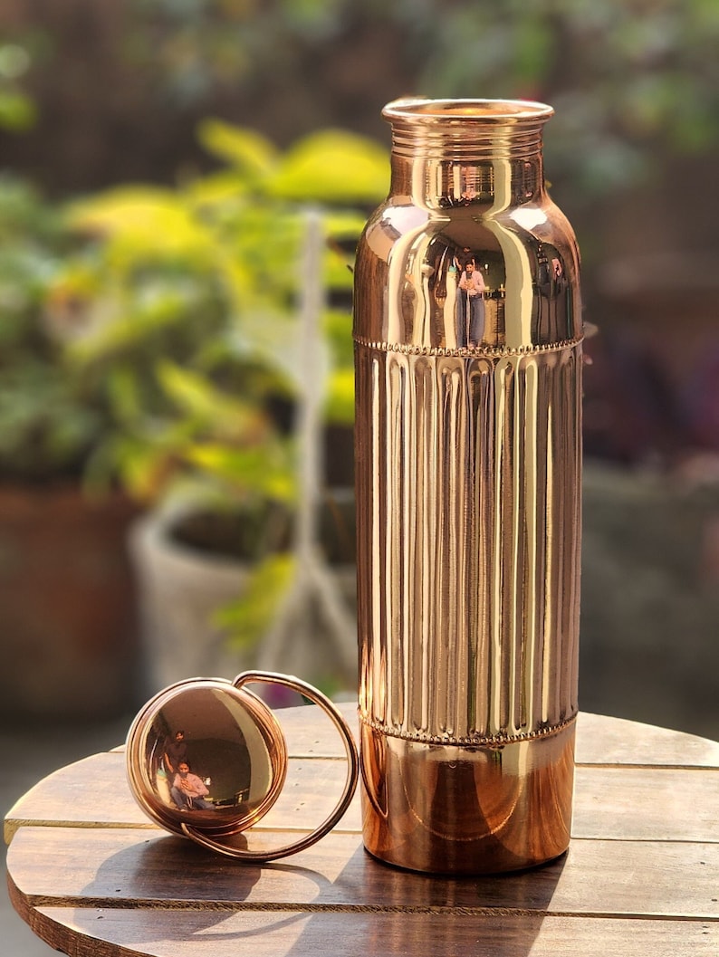 May include: A copper water bottle with a ribbed design and a matching lid. The bottle is on a wooden surface.