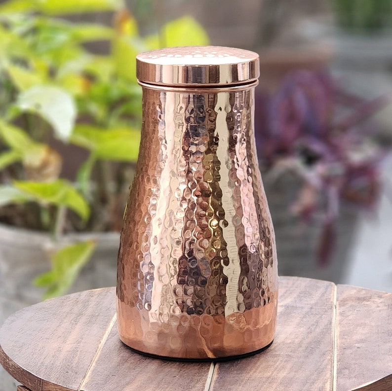 Puede incluir: Una botella de agua de color cobre con una textura martillada y una tapa a juego. La botella tiene una forma redondeada, con una base m&aacute;s ancha y un cuello m&aacute;s estrecho. El fondo est&aacute; borroso, mostrando vegetaci&oacute;n y una superficie de madera.