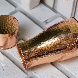 Puede incluir: Un jarr&oacute;n de metal martillado de color cobre con una tapa a juego. El jarr&oacute;n tiene una forma acampanada con una abertura ancha y un interior negro. La tapa es un peque&ntilde;o recipiente redondo con una textura martillada. Los objetos se muestran sobre una superficie de ladrillo blanco.