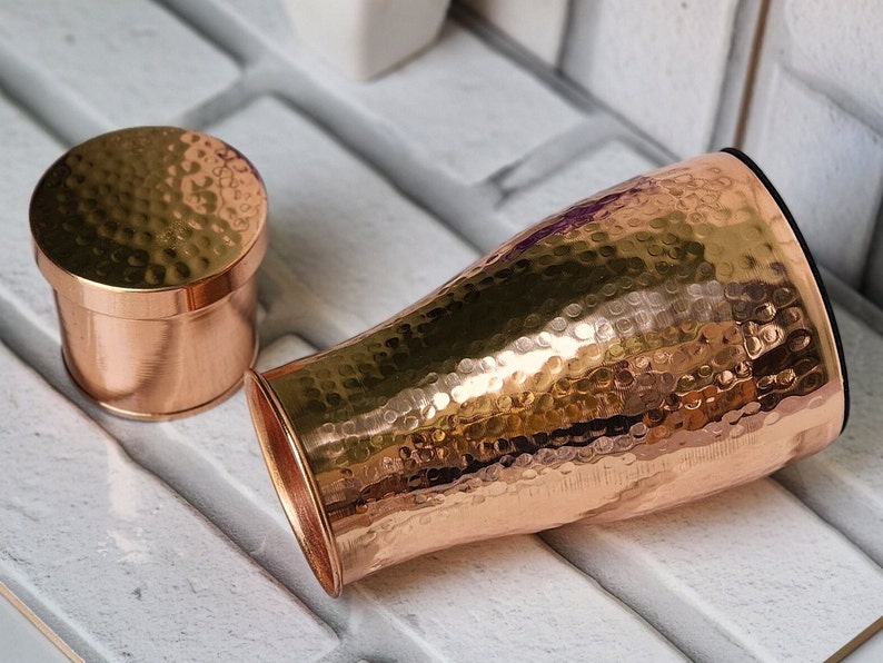 Puede incluir: Un jarr&oacute;n de metal martillado de color cobre con una tapa a juego. El jarr&oacute;n tiene una forma acampanada con una abertura ancha y un interior negro. La tapa es un peque&ntilde;o recipiente redondo con una textura martillada. Los objetos se muestran sobre una superficie de ladrillo blanco.