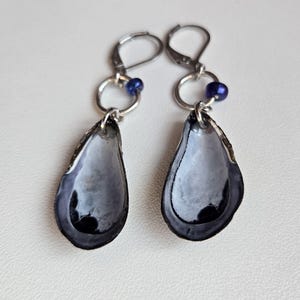 Puede incluir: Un par de pendientes hechos a mano con conchas en forma de lágrima en tonos grises y negros. Cada concha está suspendida de un cierre de palanca plateado con un pequeño acento de cuentas azules. Los pendientes están diseñados para orejas perforadas.