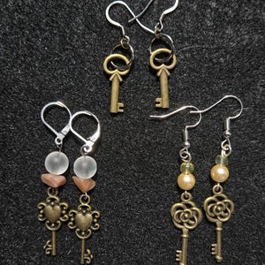 Puede incluir: Cuatro pares de pendientes con diseños de llaves y cuentas. Dos pares tienen dijes de llave con un lazo simple, un par tiene dijes de llave en forma de corazón con cuentas y el último par tiene dijes de llave en forma de flor con perlas.