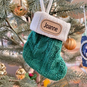 Peut inclure: Une petite chaussette de Noël tricotée verte et blanche avec le nom "Joanne" sur une étiquette en bois. La chaussette est suspendue à un sapin de Noël avec des décorations dorées et argentées.