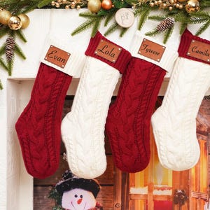 Op de afbeelding: Vier gepersonaliseerde kerstsokken hangen aan een witte schoorsteenmantel. Twee zijn rood kabelgebreid en twee zijn wit. Elke sok heeft een leren label met een naam: Evan, Lola, Tyrese en Camila. Feestelijke vakantiedecoratie.