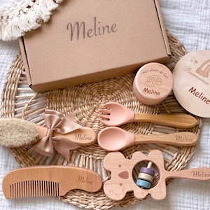 Peut inclure: Un coffret cadeau pour bébé avec des articles en bois et roses. Comprend une boîte, une brosse, un peigne, une cuillère, une fourchette, un hochet et une plaque en bois ronde. L'ensemble est personnalisé avec le nom "Meline" et d'autres textes.