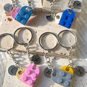 Könnte beinhalten: Schlüsselanhänger mit herzförmigen Anhängern aus bunten Bausteinen. Jeder Schlüsselanhänger hat eine silberne Kette, einen runden Initialen-Anhänger und ein geteiltes Herzdesign in Rosa, Blau, Gelb und Grau. Die Schlüsselanhänger liegen auf einer hellen Oberfläche.