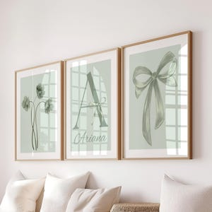May include: Three framed art prints with a pale green background. One print shows flowers, another a letter "A" with the name "Ariana", and the third a bow. All are in wooden frames.