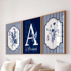 May include: Three framed wall art pieces with a floral and butterfly theme. The center piece features a large white letter "A" with the name "Ariana" below. The other two pieces have blue floral designs and butterflies.