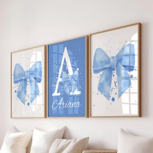 May include: Three framed wall art pieces featuring watercolor blue bows. The center frame has a blue background with a white letter "A" and the name "Ariana" in script. The other two frames have white backgrounds with blue bow designs.
