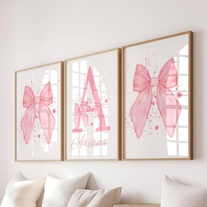 May include: Three framed art prints with a light wood finish. Each print features a watercolor-style pink bow design on a white background with pink paint splatters. The center print has the letter "A" and the name "Ariana".