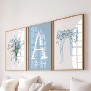 Puede incluir: Tres impresiones enmarcadas con temática floral. Las impresiones presentan un ramo de flores azules, una gran letra "A" con el nombre "Ariana" y un lazo azul con detalles florales. Los marcos tienen un acabado marrón claro.